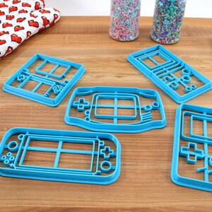 May include: Five blue plastic cookie cutters shaped like different handheld video game consoles.