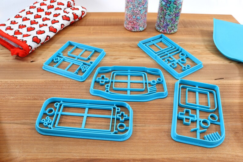 May include: Five blue plastic cookie cutters shaped like different handheld video game consoles.