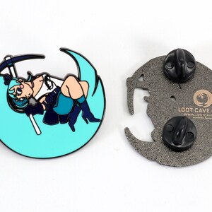 May include: A teal blue enamel pin featuring a cartoon character holding a pickaxe, sitting on a crescent moon. The back of the pin is silver with the text "Loot Cave Co.  www.lootcave.com" and two black butterfly clasps.