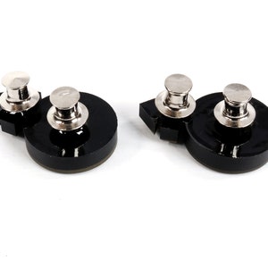 May include: Two black plastic bases with silver metal push pins. The bases are round with a slightly raised edge. The push pins are cylindrical with a flat top.