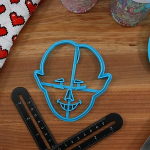 Pu&ograve; includere: Un tagliapasta in plastica blu a forma di viso con un ampio sorriso e un naso grande. Il tagliapasta &egrave; su una superficie di legno.