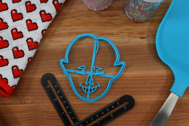 Pu&ograve; includere: Un tagliapasta in plastica blu a forma di viso con un ampio sorriso e un naso grande. Il tagliapasta &egrave; su una superficie di legno.