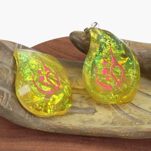 May include: Two teardrop-shaped pendants with a gold and green iridescent finish. The pendants have a pink design inside. The pendants are sitting on a wooden hand.