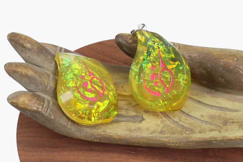 May include: Two teardrop-shaped pendants with a gold and green iridescent finish. The pendants have a pink design inside. The pendants are sitting on a wooden hand.