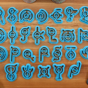 May include: A set of 24 blue plastic cookie cutters shaped like different symbols. The cutters are arranged in four rows of six on a wooden surface.