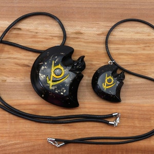 May include: Two black necklaces with gold accents. The pendants are heart-shaped with a stylized symbol in gold. The necklaces are on a wooden surface.