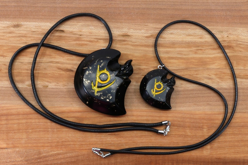May include: Two black necklaces with gold accents. The pendants are heart-shaped with a stylized symbol in gold. The necklaces are on a wooden surface.