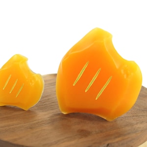 May include: Two yellow wax melts shaped like flames with three lines etched into each. The wax melts are sitting on a wooden surface.