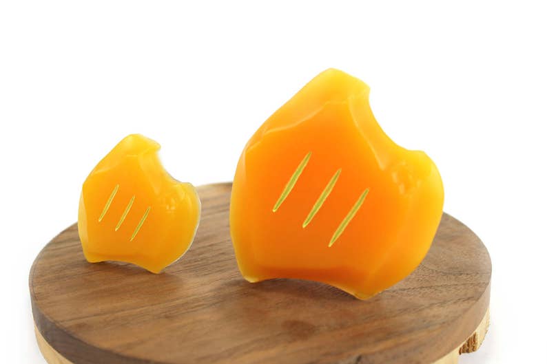 May include: Two yellow wax melts shaped like flames with three lines etched into each. The wax melts are sitting on a wooden surface.