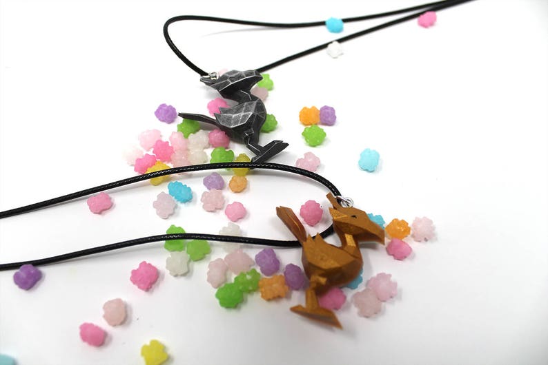 May include: Two necklaces with black cords and pendant charms. One charm is a silver-colored bird, the other is a gold-colored bird. The necklaces are surrounded by colorful candy-like sprinkles.