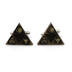 May include: A pair of gold-toned cufflinks shaped like a triangle with a stylized design inside. The design features a stylized bird, a star, a crescent moon, and a stylized animal head.