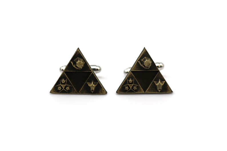 May include: A pair of gold-toned cufflinks shaped like a triangle with a stylized design inside. The design features a stylized bird, a star, a crescent moon, and a stylized animal head.