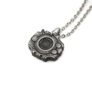 Puede incluir: Un collar de plata con un colgante con forma de dispositivo futurista. El colgante tiene una ventana circular con un cuadrado oscuro en el centro.