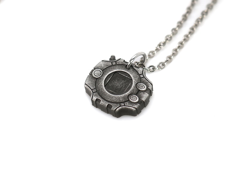 Puede incluir: Un collar de plata con un colgante con forma de dispositivo futurista. El colgante tiene una ventana circular con un cuadrado oscuro en el centro.