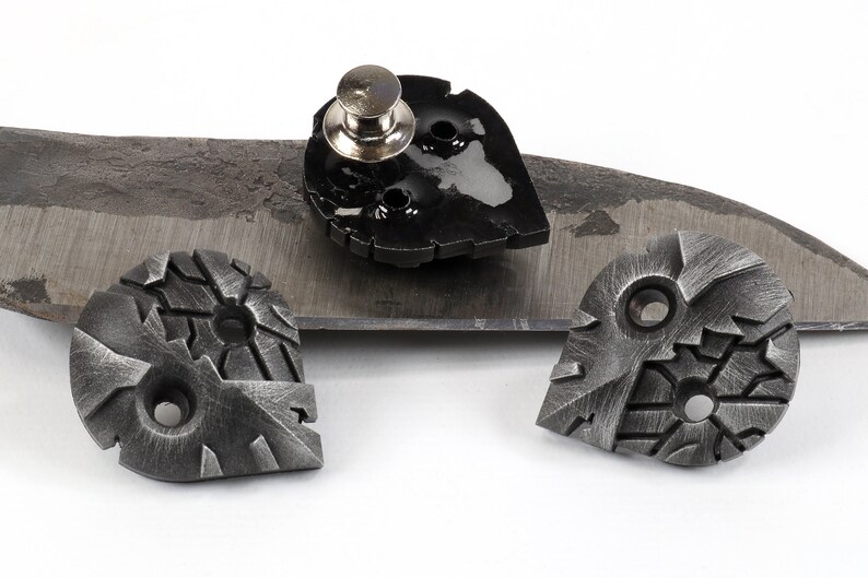 May include: Three metal heart-shaped charms with a textured surface. One charm has a silver pin attached to the back. The charms are on a gray surface.