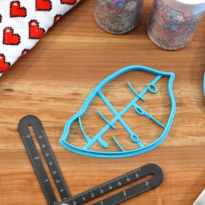 May include: A blue plastic leaf-shaped cookie cutter with a detailed vein pattern. The cutter is on a wooden surface with a ruler and a blue spatula.