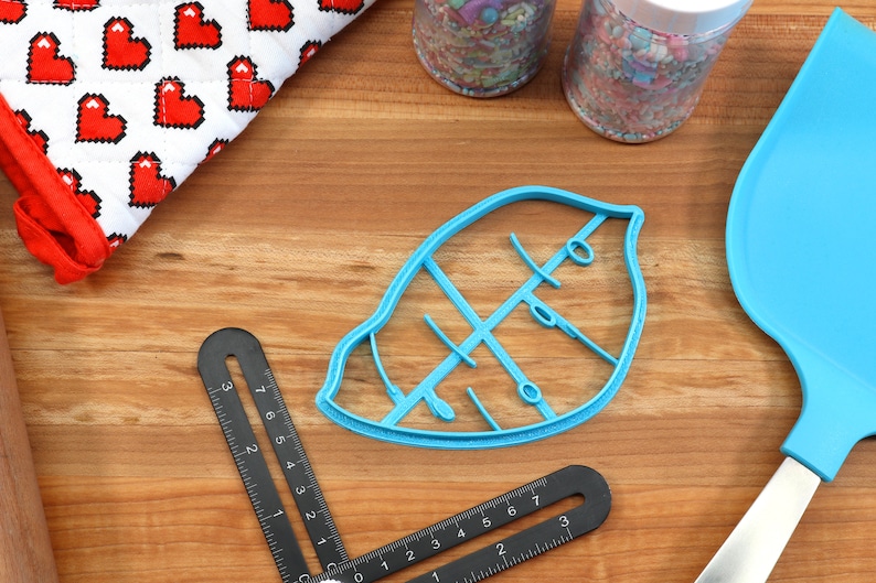 May include: A blue plastic leaf-shaped cookie cutter with a detailed vein pattern. The cutter is on a wooden surface with a ruler and a blue spatula.