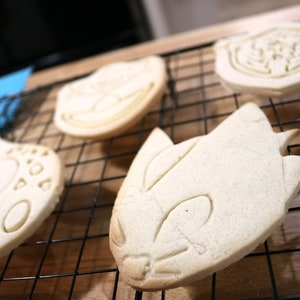 May include: A close-up of four plain sugar cookies on a black wire rack. The cookies are shaped like cartoon characters and are ready to be decorated.