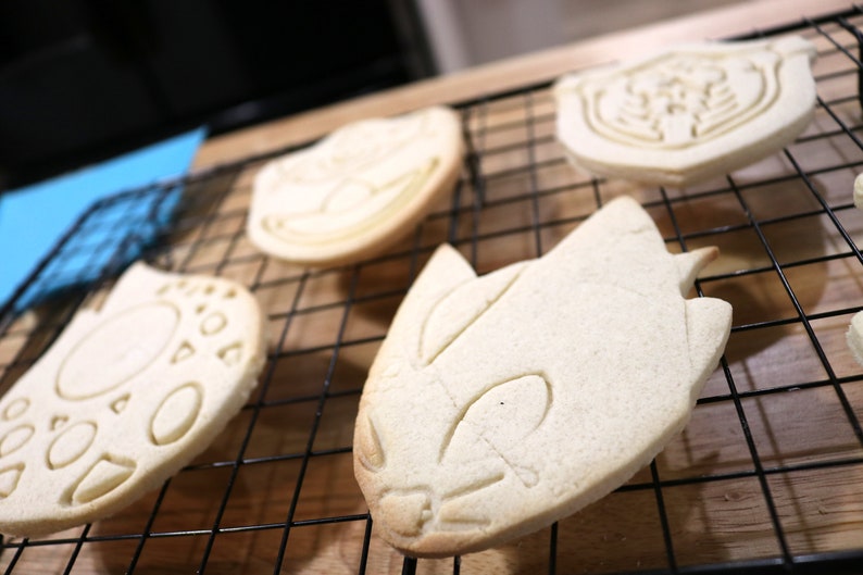 May include: A close-up of four plain sugar cookies on a black wire rack. The cookies are shaped like cartoon characters and are ready to be decorated.