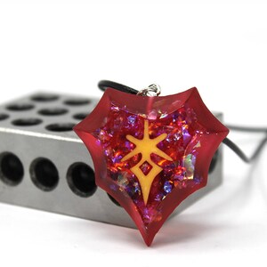 May include: A red, star-shaped pendant necklace with a yellow symbol inside. The pendant is made of resin and has a glittery finish.