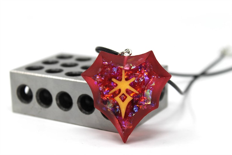 May include: A red, star-shaped pendant necklace with a yellow symbol inside. The pendant is made of resin and has a glittery finish.