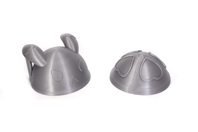 Op de afbeelding: Twee 3D-geprinte grijze plastic hoeden in de vorm van een konijn. De ene hoed is geassembleerd, de andere is gedemonteerd en laat de twee delen zien waaruit de hoed bestaat.