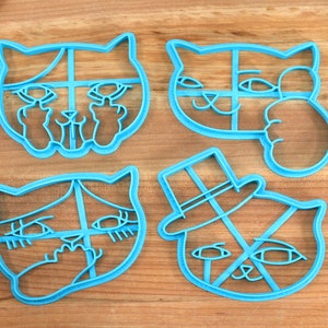 May include: Four blue plastic cookie cutters shaped like cats with different expressions. The cutters are on a wooden surface.