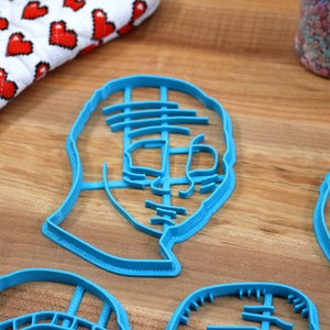 May include: Four blue plastic cookie cutters shaped like faces. The cutters are on a wooden surface.