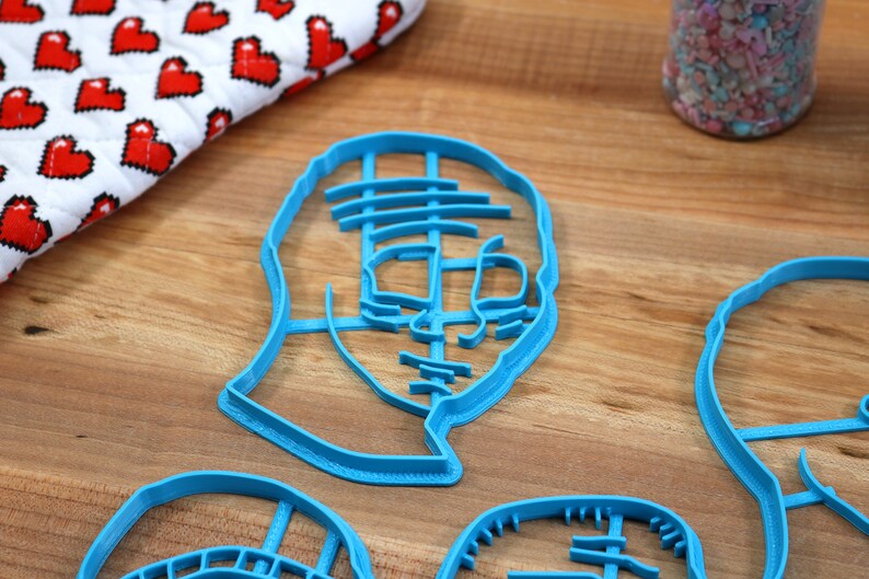 May include: Four blue plastic cookie cutters shaped like faces. The cutters are on a wooden surface.