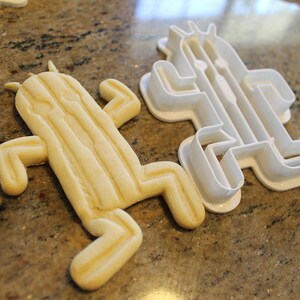 May include: A baked cactus-shaped cookie next to a white plastic cactus-shaped cookie cutter. The cookie is light yellow with vertical lines and a rounded shape. The cookie cutter is white and has a similar shape.