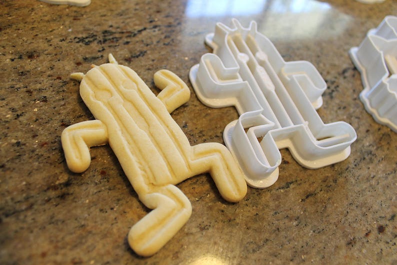 May include: A baked cactus-shaped cookie next to a white plastic cactus-shaped cookie cutter. The cookie is light yellow with vertical lines and a rounded shape. The cookie cutter is white and has a similar shape.