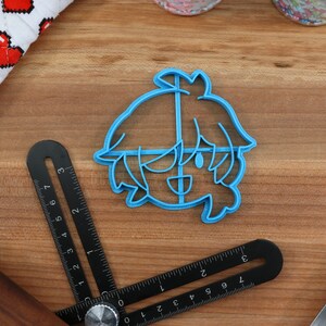 May include: A blue plastic cookie cutter shaped like a cartoon character with short, spiky hair. The character has a wide smile and large eyes.
