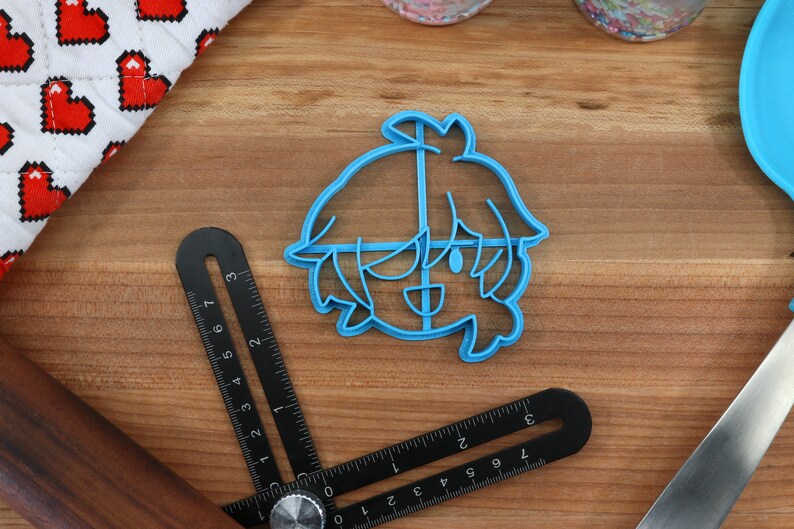 May include: A blue plastic cookie cutter shaped like a cartoon character with short, spiky hair. The character has a wide smile and large eyes.