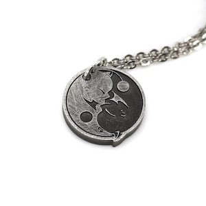 May include: A silver necklace with a yin yang pendant featuring two cartoon characters. The pendant is round and has a textured surface.