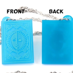 Puede incluir: Un collar de cadena plateada con un colgante azul. La parte delantera del colgante presenta un dise&ntilde;o blanco y azul de una cara con un sol radiante a su alrededor. La parte posterior del colgante es de color azul s&oacute;lido.