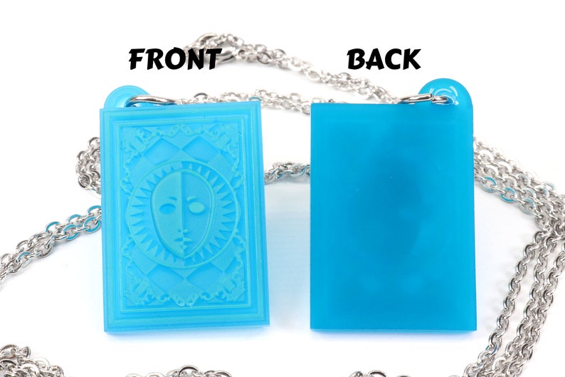 Puede incluir: Un collar de cadena plateada con un colgante azul. La parte delantera del colgante presenta un dise&ntilde;o blanco y azul de una cara con un sol radiante a su alrededor. La parte posterior del colgante es de color azul s&oacute;lido.