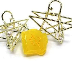 May include: Two gold-colored star-shaped hair clips with a pearl accent. A yellow, irregularly shaped object with a circular design is between the clips. The clips have a geometric design.