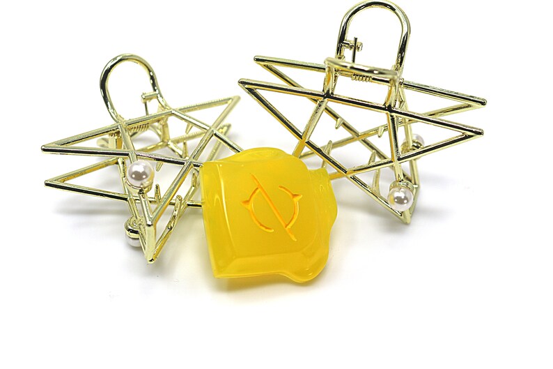 May include: Two gold-colored star-shaped hair clips with a pearl accent. A yellow, irregularly shaped object with a circular design is between the clips. The clips have a geometric design.