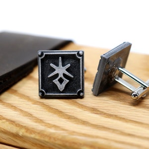 May include: A pair of silver cufflinks with a black square design featuring a stylized symbol. The cufflinks are on a wooden surface.