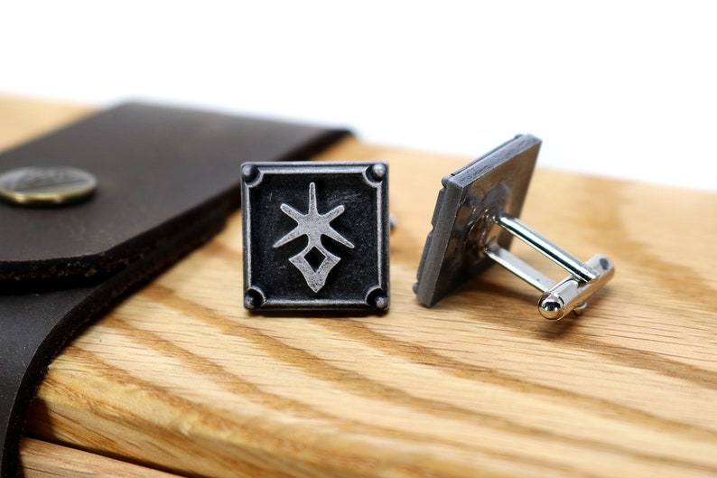 May include: A pair of silver cufflinks with a black square design featuring a stylized symbol. The cufflinks are on a wooden surface.