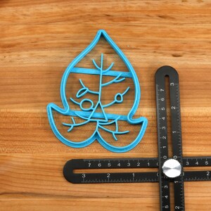 May include: A blue plastic cookie cutter shaped like a leaf with a detailed design. The cutter is on a wooden surface with a ruler next to it.