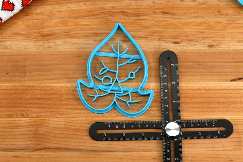 May include: A blue plastic cookie cutter shaped like a leaf with a detailed design. The cutter is on a wooden surface with a ruler next to it.