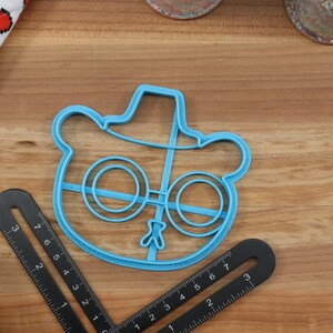 May include: Blue plastic cookie cutter shaped like a cartoon bear wearing a hat and glasses.