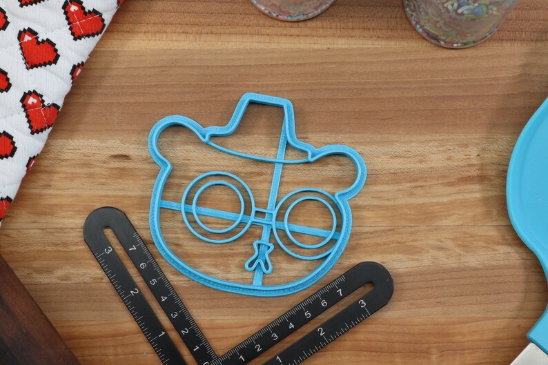 May include: Blue plastic cookie cutter shaped like a cartoon bear wearing a hat and glasses.