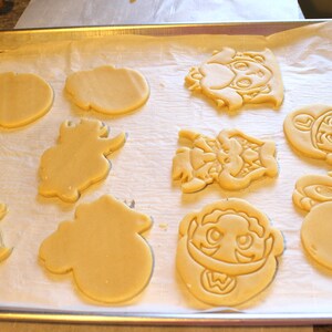 May include: A baking sheet with unbaked sugar cookies cut into various shapes, including a mushroom, a star, a dinosaur, and a character from a video game.