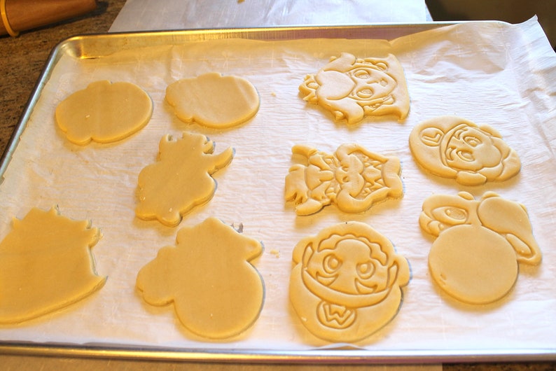 May include: A baking sheet with unbaked sugar cookies cut into various shapes, including a mushroom, a star, a dinosaur, and a character from a video game.