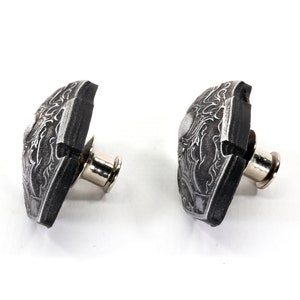 May include: Two black and silver bicycle toe clips with a decorative design. The toe clips are attached to a metal post.