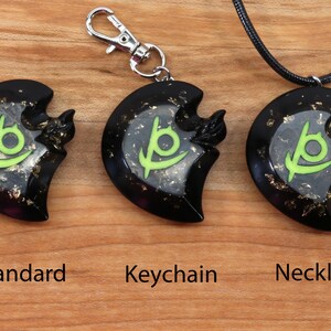 May include: Three black crescent moon pendants with a green symbol inside. The pendant on the left is a standard pendant, the middle pendant is attached to a silver keychain, and the right pendant is attached to a black cord necklace.