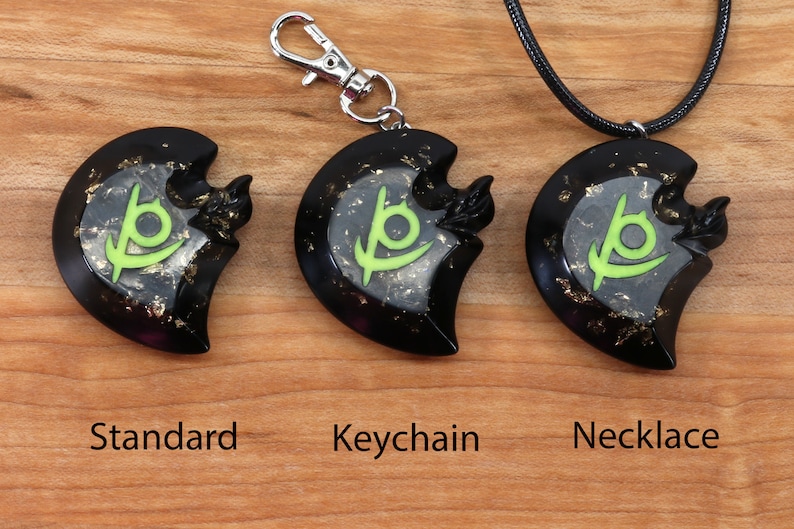 May include: Three black crescent moon pendants with a green symbol inside. The pendant on the left is a standard pendant, the middle pendant is attached to a silver keychain, and the right pendant is attached to a black cord necklace.