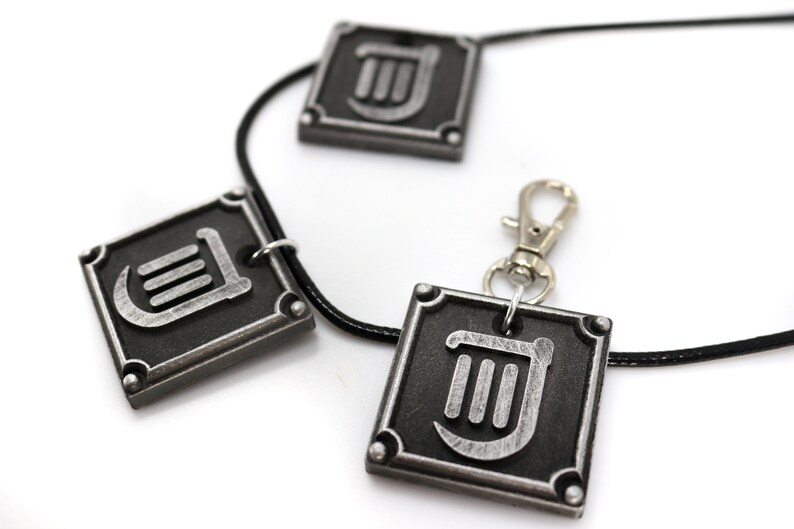 May include: A black leather necklace with three silver pendants featuring a stylized lyre symbol. The pendants are square shaped with rounded corners and have a silver finish.